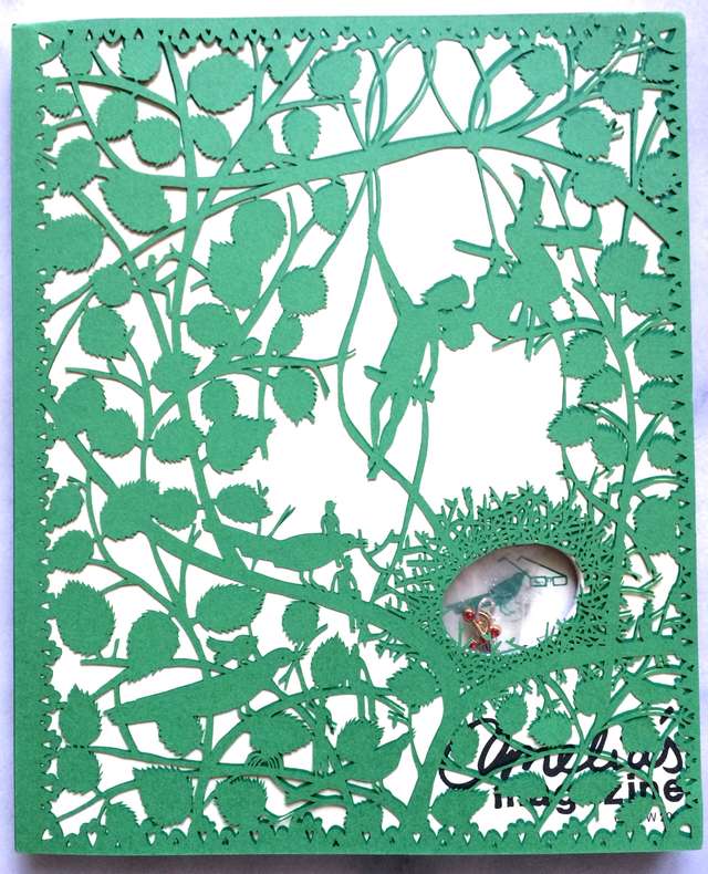 A magazine with an intricate cut out green cover suggesting a thicket, fairies and real treasure stuck in a hole cut into the magazine.