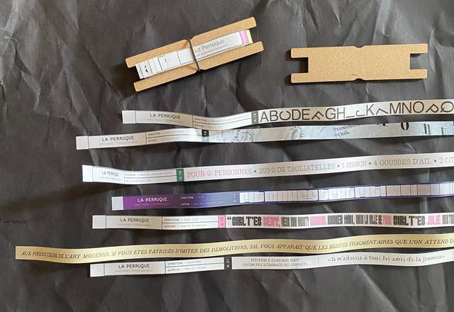 Strips of paper that show examples of various type faces. Above them are 2 card winders, one full with the strips wound around it and secured, the other empty.
